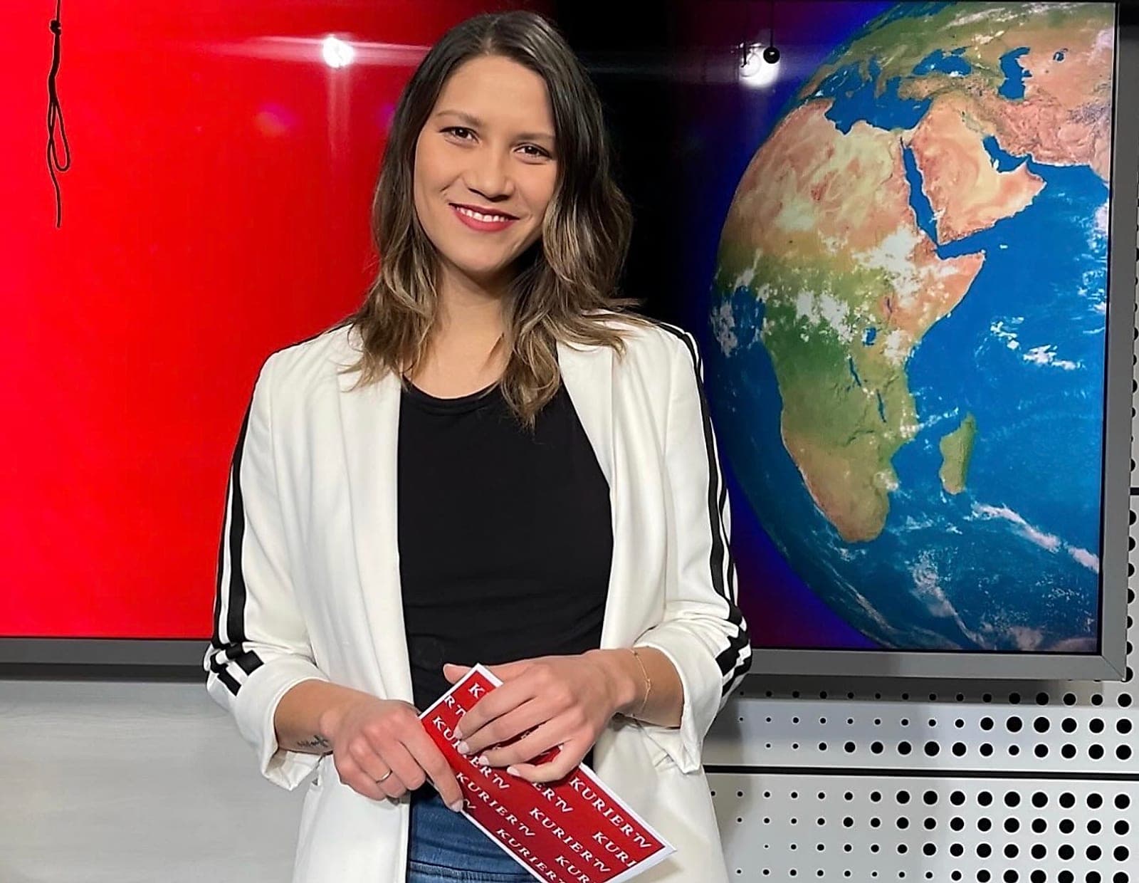 Judith Tieber-Gibbs im Fernsehstudio, vor einer roten Wand und einem Globus auf dem Bildschirm.