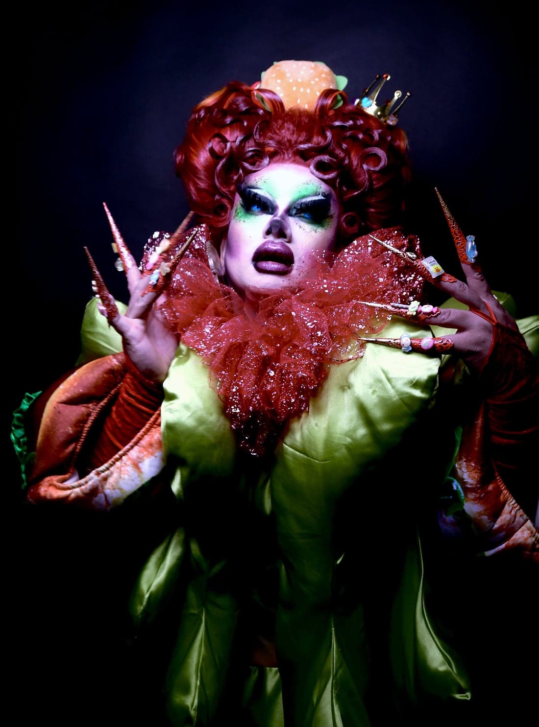 Crystal O in the studio, red curls with a burger crown, a glittering ruff and a green satin gown, with extra long nails.