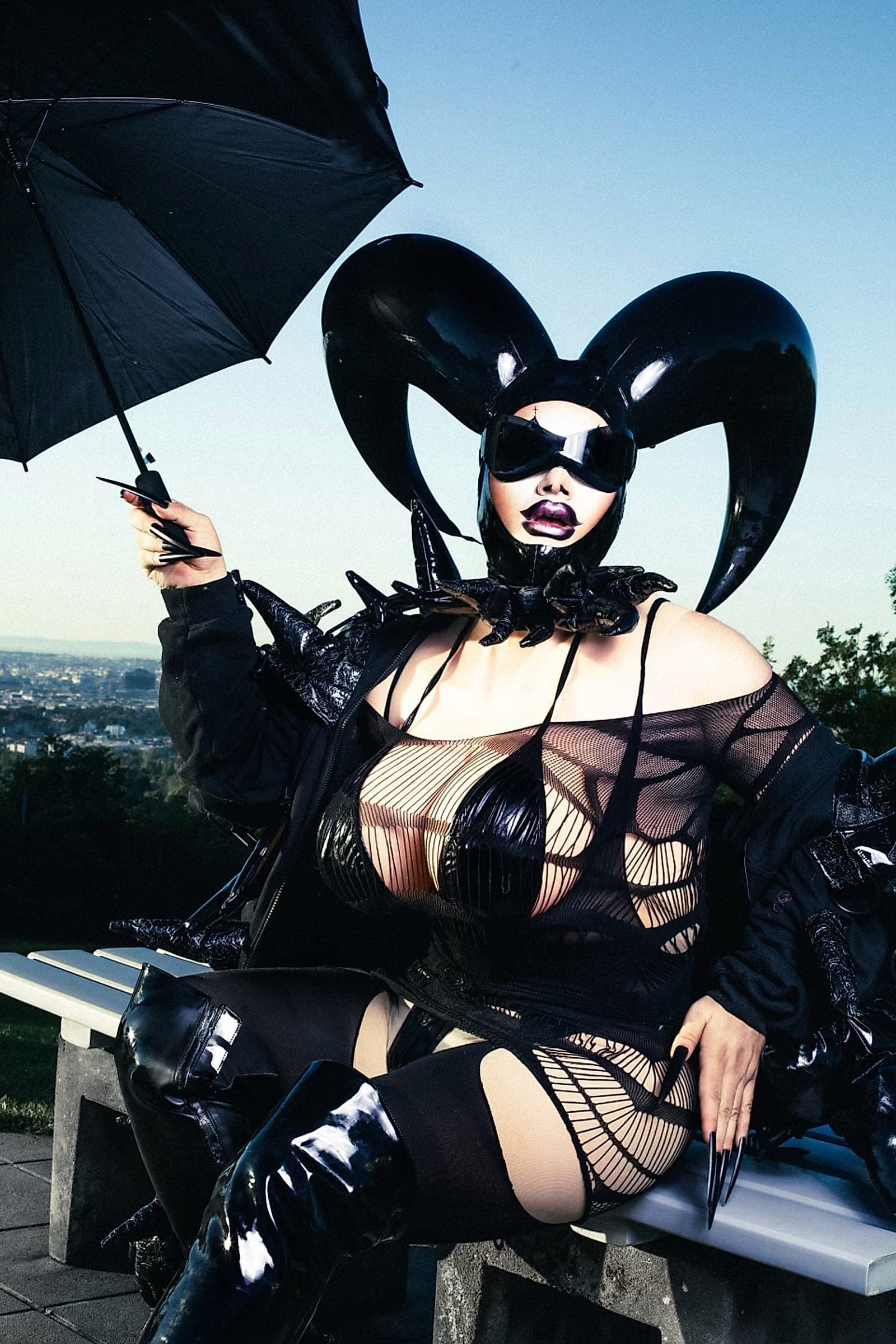 Crystal O in a black latex outfit with a curved headpiece, holding an umbrella, against the sky above Vienna.