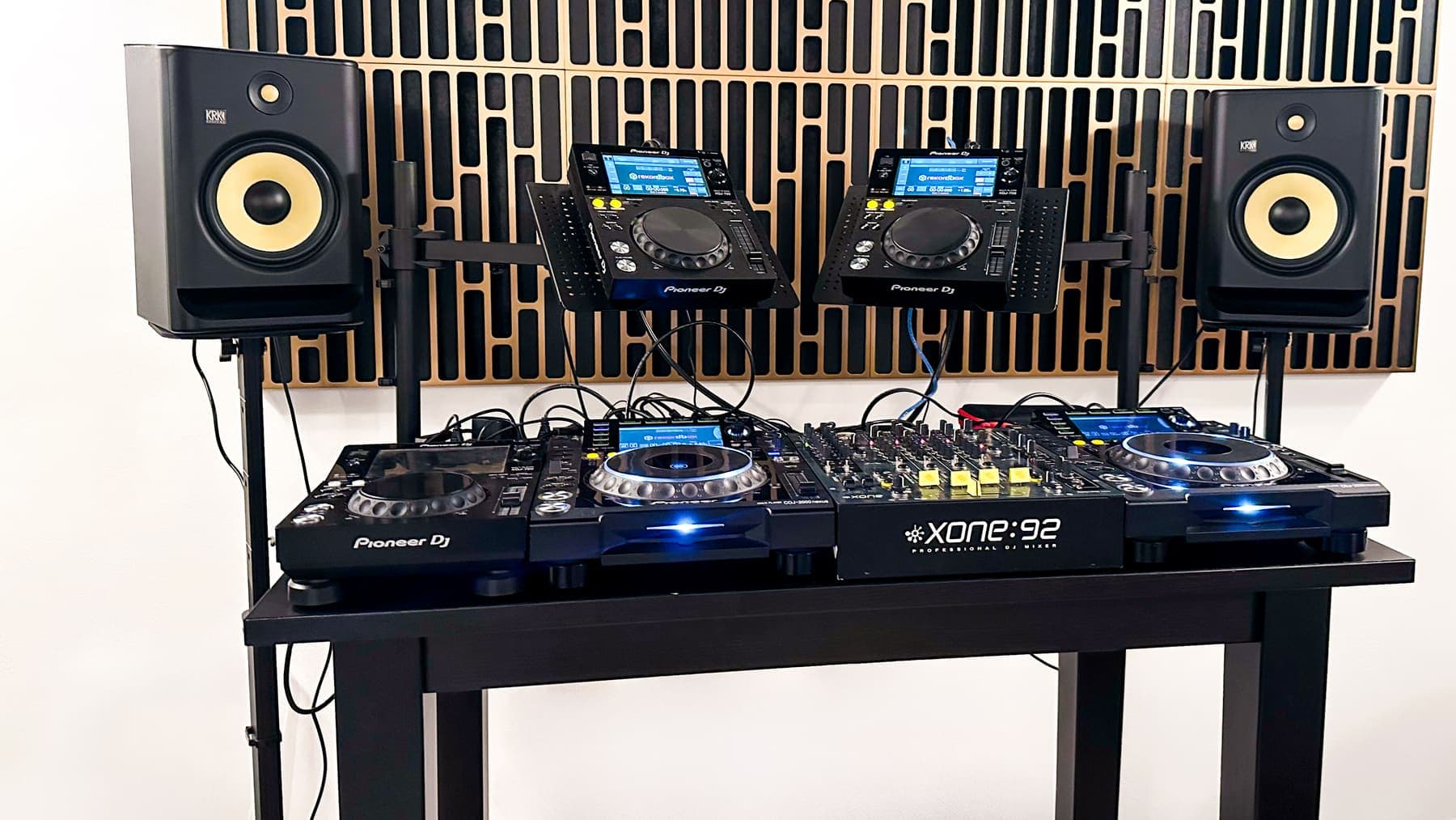 DJ setup in the studio: two players and a mixer on a table, with two studio monitors and a slatted wood wall behind them.