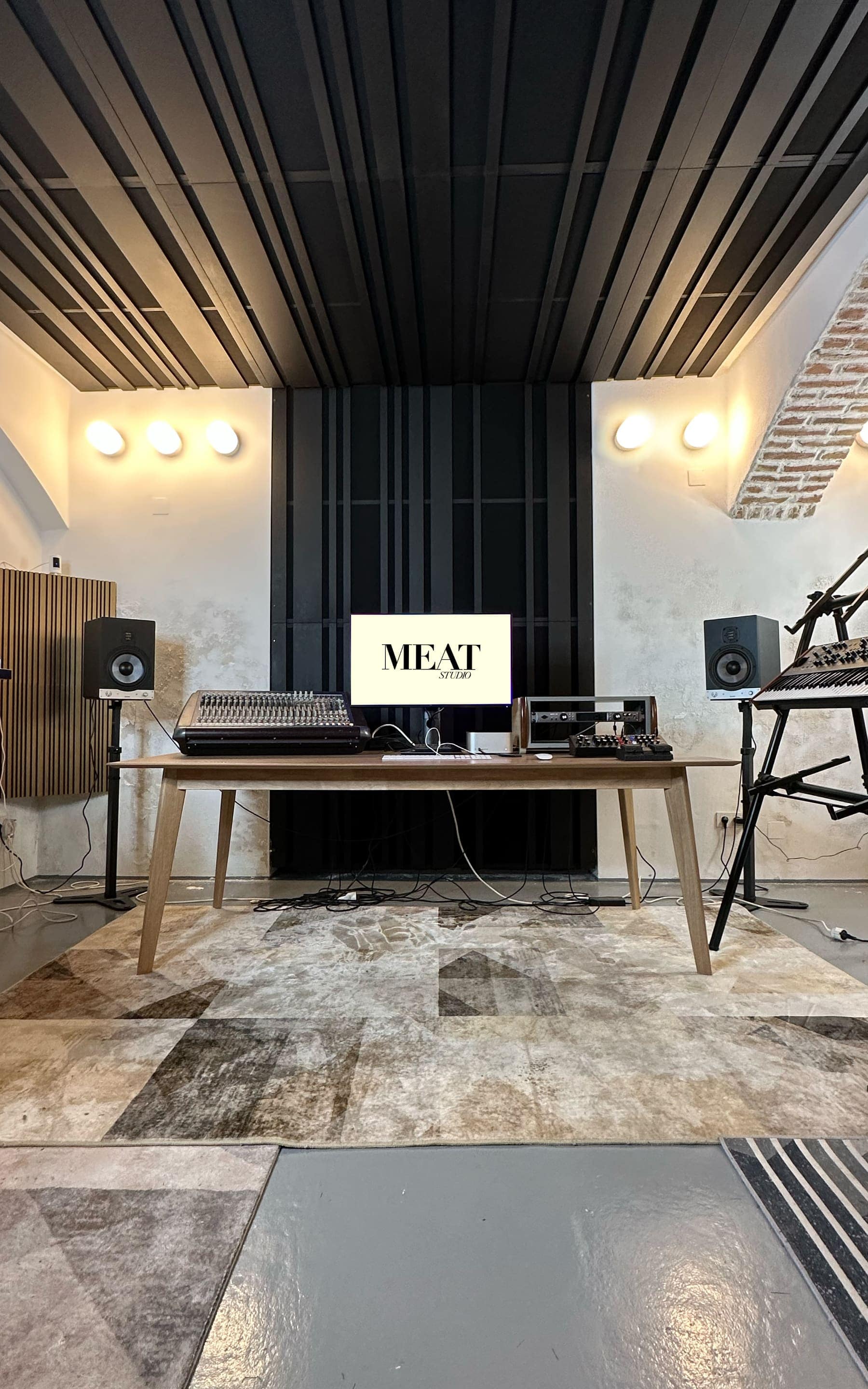 The studio room from the front: a long wooden table with mixing desk and screen, studio monitors left and right, black wooden slats on the ceiling.