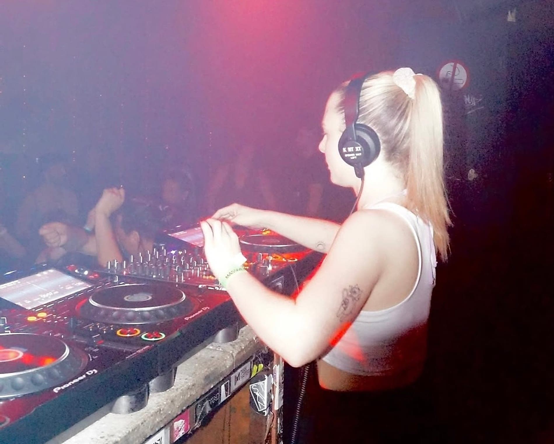 Violetta playing a club set, headphones on one ear, hands on the mixer, the crowd behind her in red light.