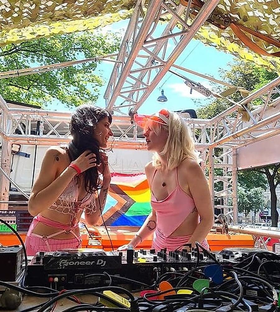Annika Stein and Wered Fuchs together at the mixer during an open-air set, the Progress Pride flag behind them.